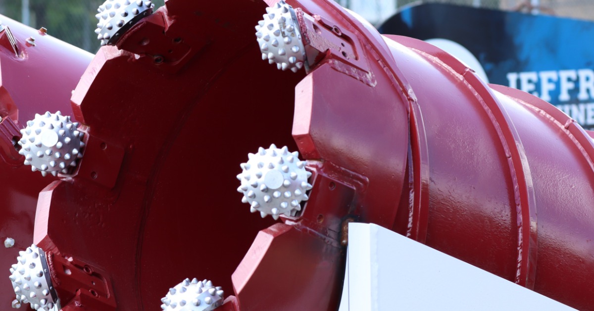 A red cylindrical drilling attachment with white spiked cutting teeth sits near a sign and other equipment outdoors.